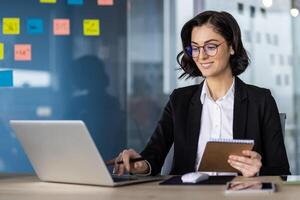 Confident businesswoman in black suit uses laptop for teamwork on project. Smiling while holding notebook in office with colorful sticky notes. Engaged in online business communication. photo