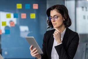 Businesswoman with glasses using a tablet in office environment, surrounded by colorful sticky notes. Depicts professional engagement and technology in workspace with serious focus and concentration. photo