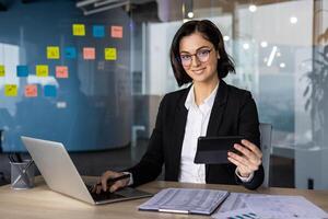 Confident businesswoman using laptop and smartphone for reports in office. Professional setting conveys productivity and efficiency in analysis and communication with technology. photo