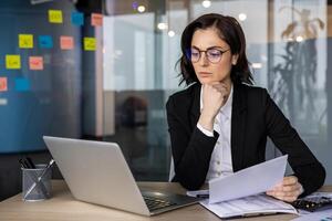 Focused businesswoman analyzing documents at office desk, laptop open alongside. Concept of concentration, analysis, and professional life. Workspace features colorful notes in background. photo