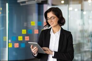 Confident professional woman in business suit uses digital tablet in modern office setting. Surrounded by colorful sticky notes on glass wall, focusing on project management and productivity. photo