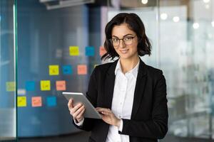 Confident businesswoman using tablet in modern office setting, engaging with tasks efficiently. Brightly colored sticky notes in background highlight teamwork and innovative ideas. photo