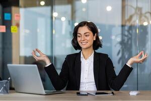 Businesswoman meditating in office at desk practicing mindfulness. She uses laptop for work tasks, creating calm focus and balance amidst busy workplace. Professional setting emphasizes career. photo