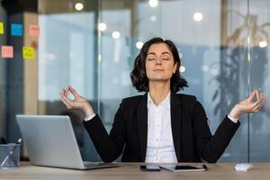 Woman in business attire meditating at office desk, eyes closed, embracing tranquility and mindfulness. Laptop and phone nearby emphasize balance between work and relaxation amidst workplace stress. photo