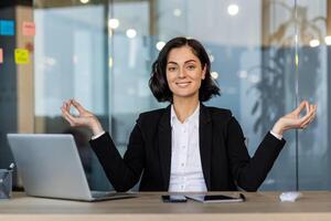 Confident woman in business suit practicing mindfulness yoga pose at office desk. Promotes calmness and focus during work. Ideal for themes related to meditation, stress management, and wellness. photo