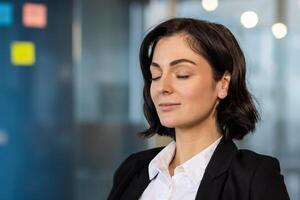 Confident businesswoman practicing mindfulness in office, eyes closed, demonstrating stress relief and inner peace in modern professional environment. Focuses on relaxation and mental well-being. photo