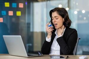 Businesswoman in office uses an inhaler for asthma relief while working at laptop. Concept of managing health at work. She is dressed in formal attire. Health and business combine. photo