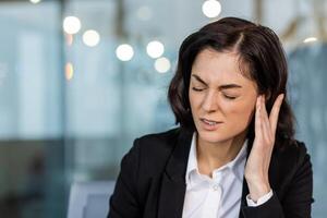 Woman at work experiencing ear pain, showing expression of discomfort. Dressed in formal business attire in office setting. Image conveys stress, possible hearing trouble, and workplace tension. photo
