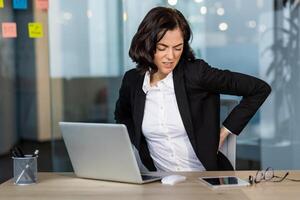 Businesswoman in office showing signs of back pain while sitting at desk using laptop and phone. Indicates discomfort, poor posture, need for ergonomic solutions in workplace. photo