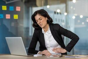 Woman in office experiencing discomfort while using laptop. Signs of stress or stomach pain. Formal business attire suggests professional setting. Expression indicates need for relief. photo