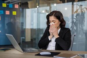 Businesswoman sneezing at office desk with laptop open, showing symptoms of cold. Captures concept of illness at work, stress, and need for better health management during work hours. photo