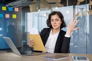Young businesswoman holding envelope in modern office, expressing confusion while discussing financial documents. Laptop and smartphone visible, creating sense of corporate frustration. photo