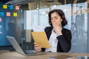 Businesswoman in modern office receiving important mail, expressing concern. Glancing at laptop, reflecting serious business communication or unexpected announcement. photo