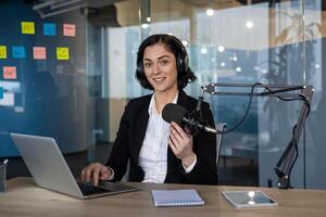 Confident podcast host in office setting, engaging with microphone. Wearing headphones, using laptop, smartphone nearby. notes display creative workspace environment, modern communication photo