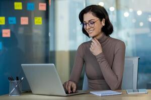 Businesswoman wearing glasses using laptop in office setting. Confidently working on tasks, showing focus and determination. Modern workspace features desk with notebook photo