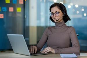 Smiling woman with glasses and headset uses laptop in office setting. Engaged in online meeting or support call. Modern technology enhances communication and productivity in business environment. photo