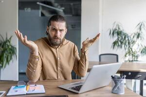 A man with a puzzled expression sits at a desk using a laptop, gesturing with raised arms, conveying confusion or hesitation while working in an office or professional setting. photo
