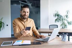 A bearded man sits at a desk in a contemporary office space, utilizing a smartphone and a laptop. Various tools and notes are present symbolizing efficiency, work, and communication. photo