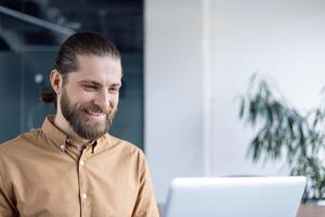 A cheerful man with a beard uses his laptop while seated in a modern office setting. The image conveys a positive mood, focusing on productivity and workplace happiness. photo