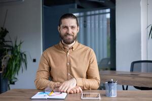 Male professional seated at desk, smiling confidently with documents and devices on the surface, conveying productivity and modernity in workplace setting. photo