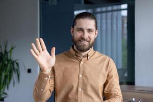 Friendly man wearing a brown shirt waves towards the camera, expressing positivity and approachability, situated in a modern office environment. photo