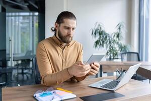 A professional man sits at a desk in a modern office, holding a tablet and focusing on work. His setup includes a laptop, notebook, and accessories indicative of productivity. photo
