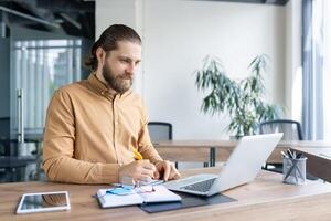 A young professional in a well-lit office working at a laptop, taking notes, exemplifying focus and productivity. The image highlights a modern workspace and portrays concentration photo