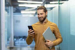 A professional man stands in a modern office setting, smiling at his smartphone while holding a laptop. The image conveys productivity and a positive workplace atmosphere. photo