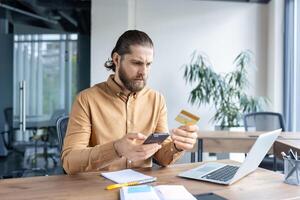 Focused man sitting at a desk in an office environment interacting with a smartphone and a credit card, implying online payment, banking, or identity verification. photo