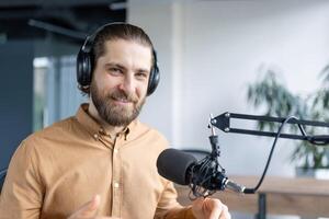 A male podcast host seated at a desk, wearing headphones and speaking into a professional microphone in a studio setup. He is smiling and exuding a confident photo