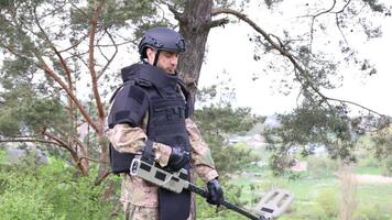 Man in a military uniform and bulletproof vest works in forest with a metal detector video
