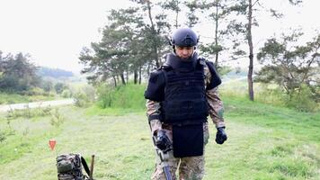 Man in a military uniform and bulletproof vest works in the forest with a metal detector. video