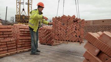 Worker guiding a crane lifting a pallet of bricks, A construction worker in safety gear guiding a pallet of bricks lifted by a crane. video