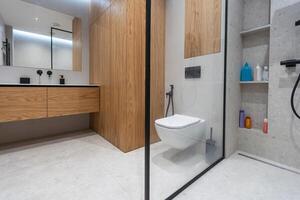 Bathroom in a modern style with gray and white tiles. There is a large mirror with luminous lamps, tabletop with wooden drawers and sink, bath with shower and glass partition, towel rack and a hanger. photo