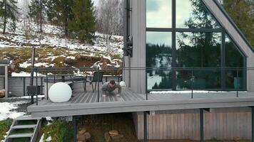 Man exercising on a wooden terrace, in front of a modern A-frame house with large glass windows. The terrace overlooks a snowy hillside with scattered trees and a scenic landscape video