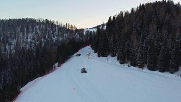 Two snowcats preparing ski slopes. Aerial view of the two vehicles between trees in the evening in winter video