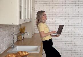 Confident latin female remote employee freelancer sit at kitchen table search files on phone to forward on laptop via home wireless network. Smart young lady user sync restore data using two gadgets photo