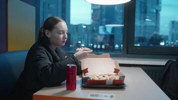 Teenage girl enjoying pizza and coke while sitting at table. Female is relaxing by window with view of city. She is wearing jacket in hotel. video