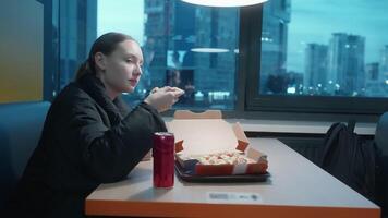 Teen girl enjoying a pizza dinner in a cafe, savoring slices and sipping a drink against the backdrop of a city nightscape viewed through a large window video
