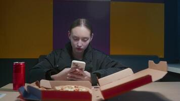 Teenage girl enjoying pizza and using a smartphone in a cafe, reflecting modern lifestyle and digital addiction amidst colorful surroundings video
