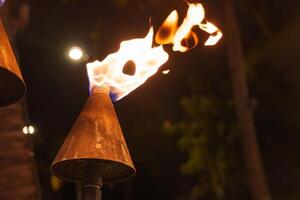A lit tiki torch with a conical metal top emits a warm glow. Blurred foliage and faint lights in the background suggest an outdoor tropical setting. photo