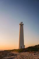 A tall white lighthouse stands against a clear sunset sky, surrounded by greenery and sandy terrain, with the ocean visible in the background. photo