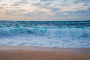 A tranquil beach scene with turquoise ocean waves, white foam, and a pastel colored sky. Soft light and clouds suggest a tropical coastal setting. photo
