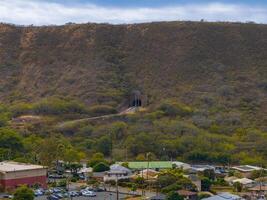 A hillside with a tunnel entrance surrounded by greenery, power lines, and a residential area with houses, parking lots, and larger buildings on Oahu. photo