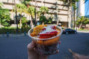 A colorful acai bowl with strawberries, mango, coconut, and granola is held against a modern building, palm trees, and a street in Honolulu. photo