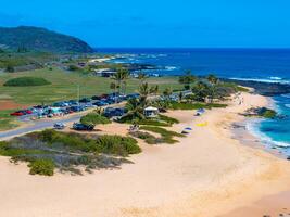 Aerial view of a sandy beach on Oahu island, Hawaii, with turquoise waters, a winding road, palm trees, and a lush green landscape in the background. photo