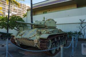 A weathered green military tank with rusted tracks is displayed outdoors near a light colored building, palm trees, and a multi story structure in Hawaii. photo