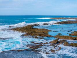 Rugged rocky formations surrounded by vibrant blue ocean waters on Oahu island. Waves crash, creating foam, with people visible near calmer pools. photo