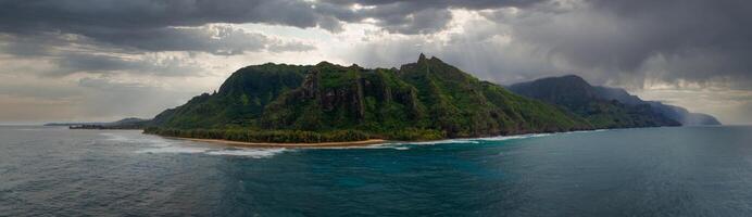 Lush green cliffs of the Napali Coast rise from the ocean, with a sandy beach and palm trees along the shoreline under a cloudy, sunlit sky. photo
