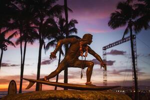 A bronze surfer statue adorned with flower leis stands against a vibrant sunset sky, with silhouetted palm trees adding to the tropical setting. photo
